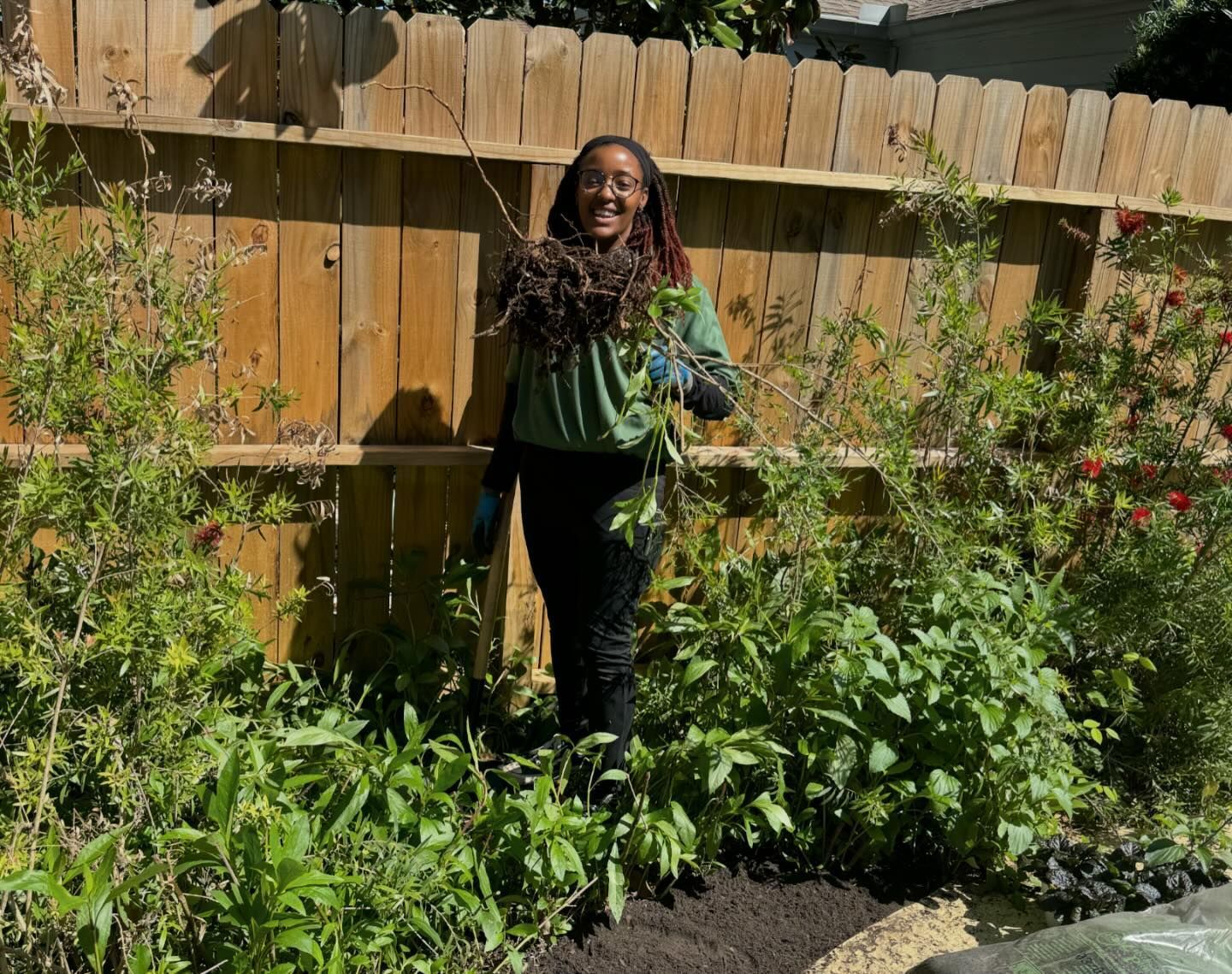 Person in garden holding branches, smiling. Wooden fence background, surrounded by green plants. Sunny day.