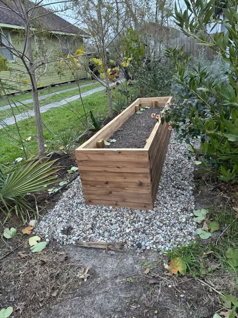 Wooden raised garden bed filled with soil on a gravel base in a yard.