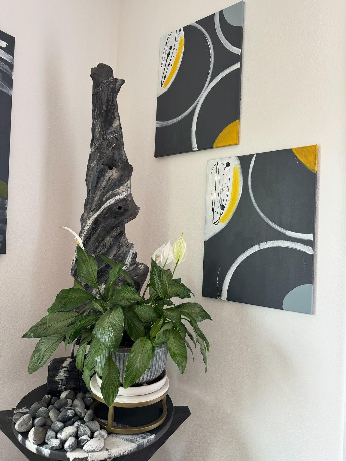 Corner decor: art with gray, yellow circles, wooden sculpture, potted plant, and black tray with stones.