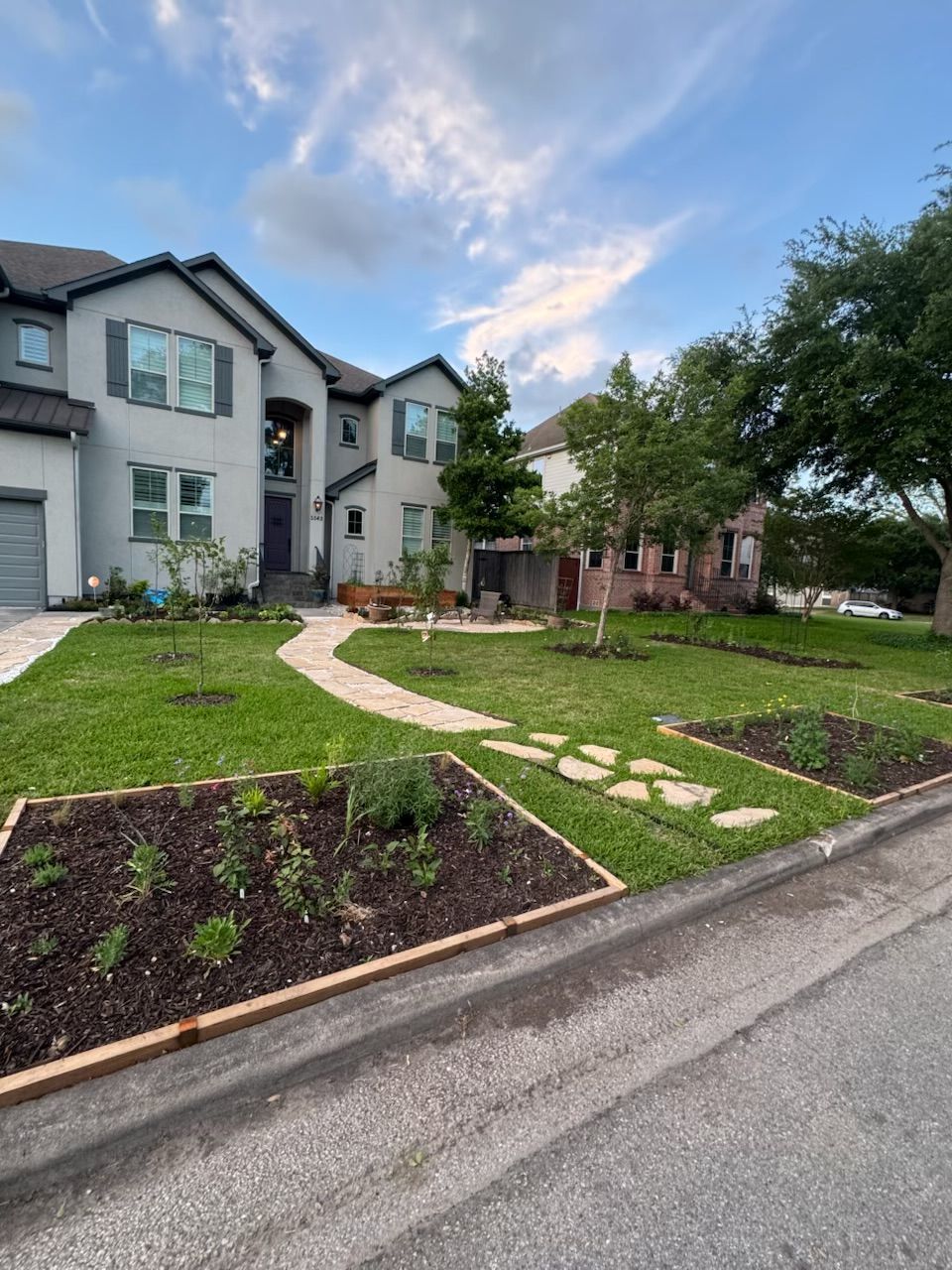 Two-story gray house with new landscaping, stone path, flower beds, and green lawn. Cloudy blue sky.