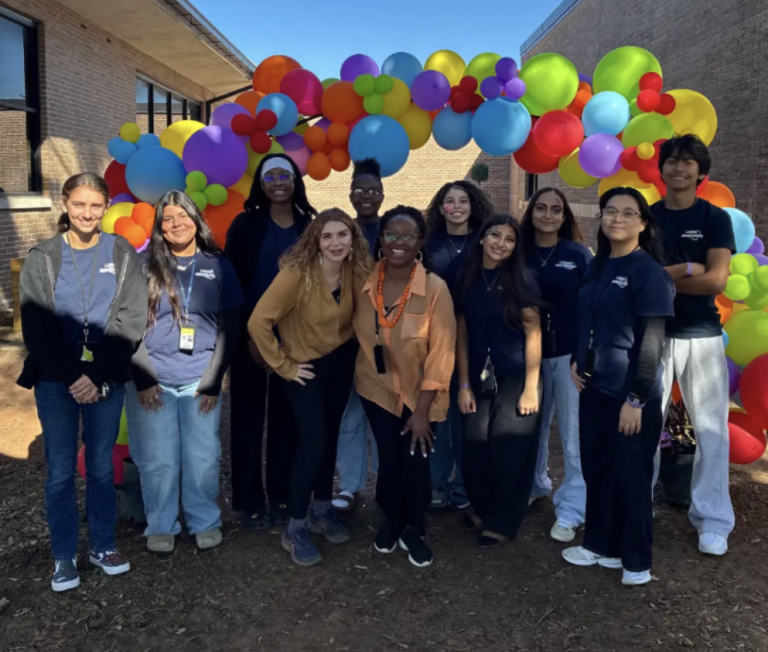 Group of people from Rooted In Sunshine standing under a colorful balloon arch outdoors, smiling at the camera.