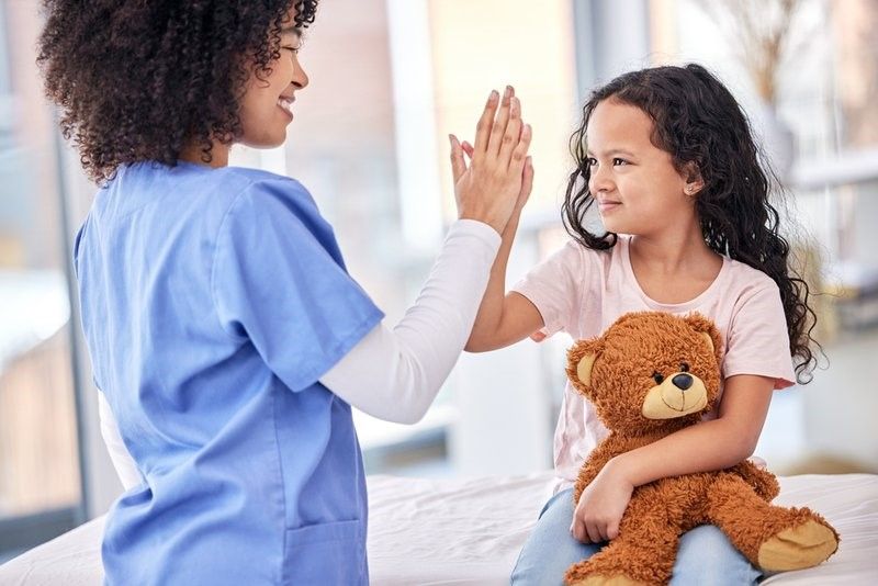 A nurse is giving a little girl a high five while holding a teddy bear. A nurse is giving a little girl a high five while holding a teddy bear.