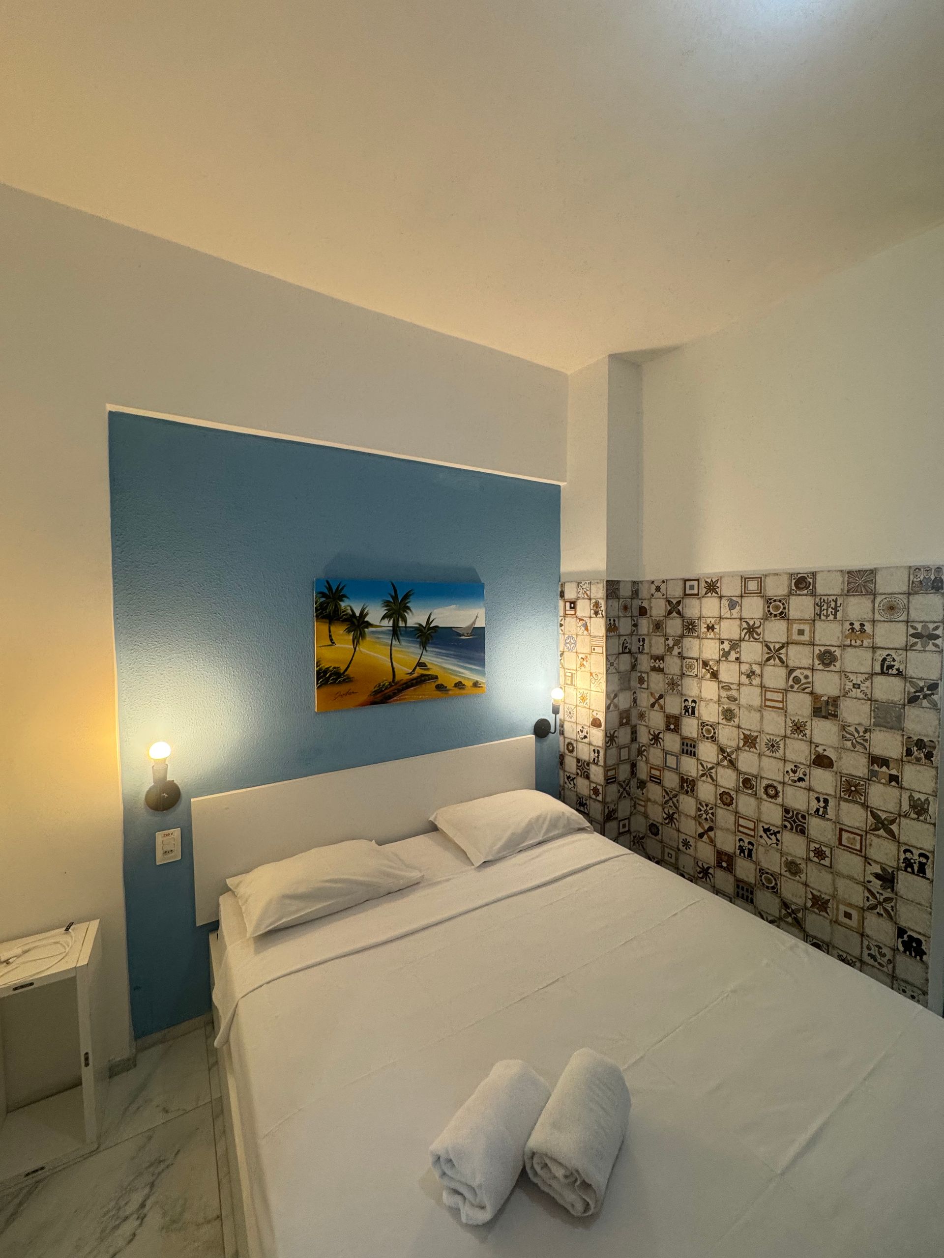 Quarto de hotel pequeno com cama branca, parede com detalhes em azul, azulejos estampados e um quadro com tema de praia.