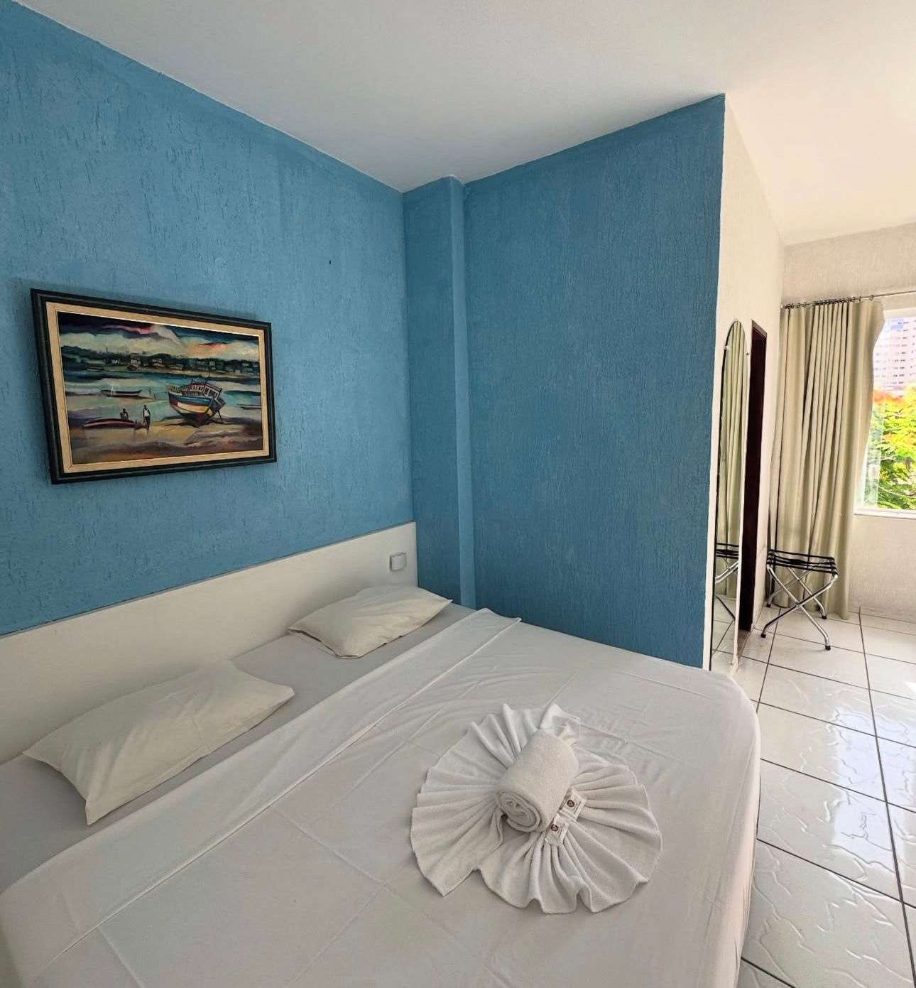 Quarto azul claro com roupa de cama branca, obras de arte emolduradas na parede e corredor com azulejos que leva a uma janela.