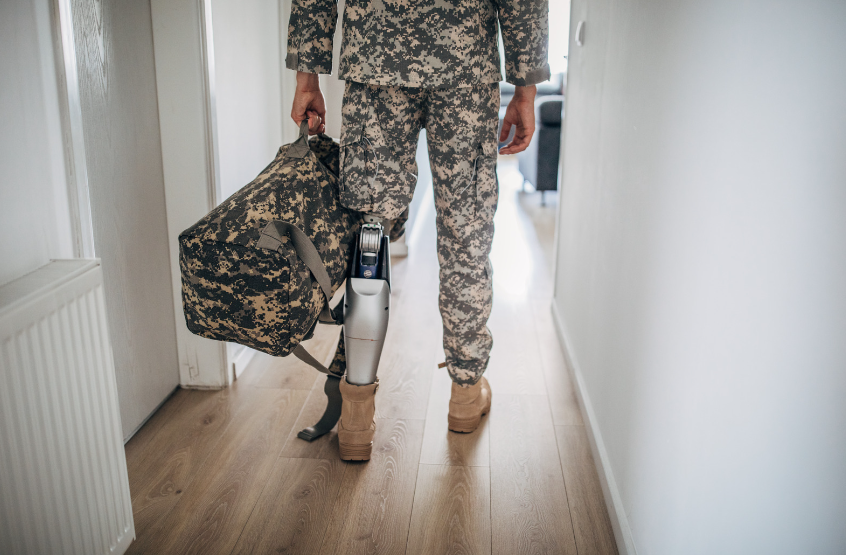 A soldier with a prosthetic leg is walking down a hallway holding a backpack.