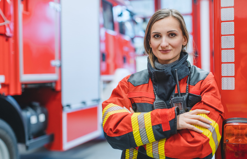 A female firefighter is standing in front of a fire truck with her arms crossed.