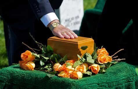 Person's hand on a wooden cremation urn surrounded by orange roses on a green cloth, near a gravestone outdoors.