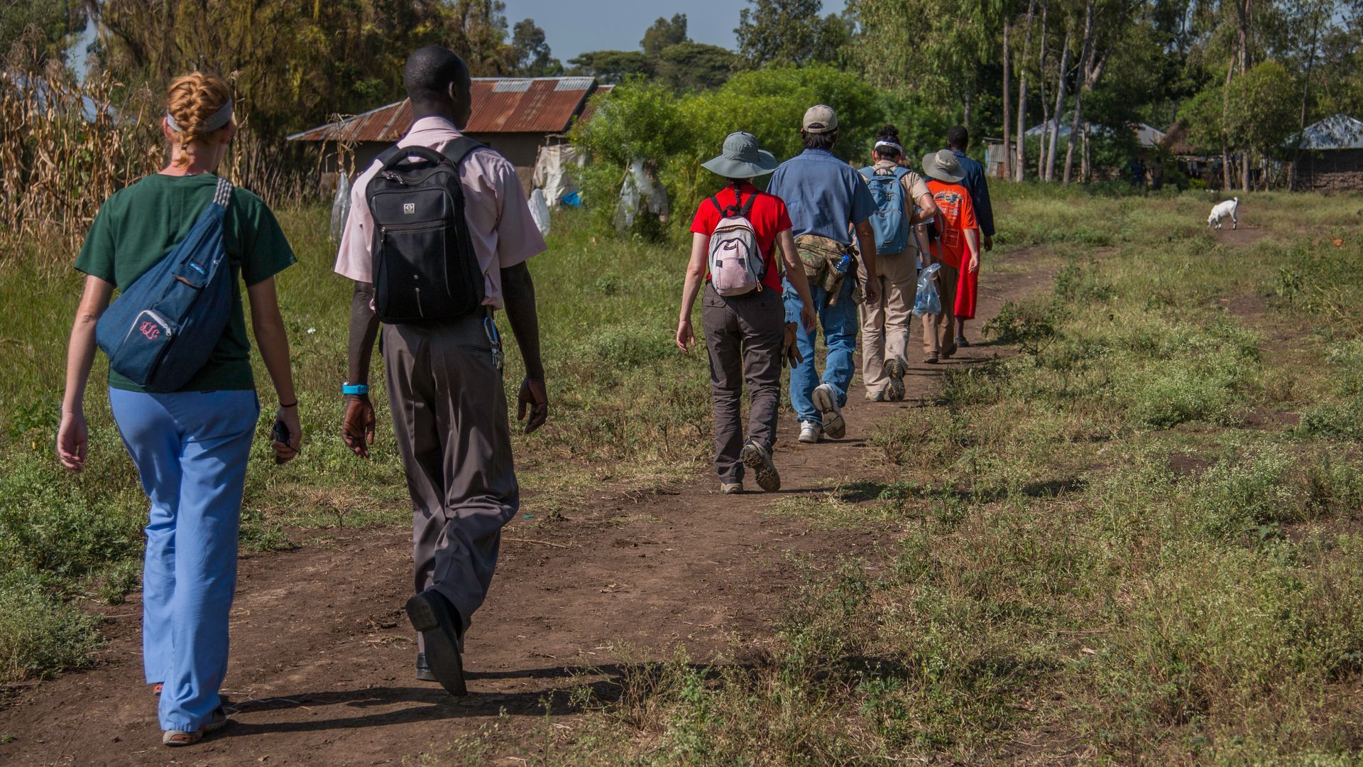 A group of people wearing casual clothing and backpacks walk together on a dirt path in a rural, sunny setting.