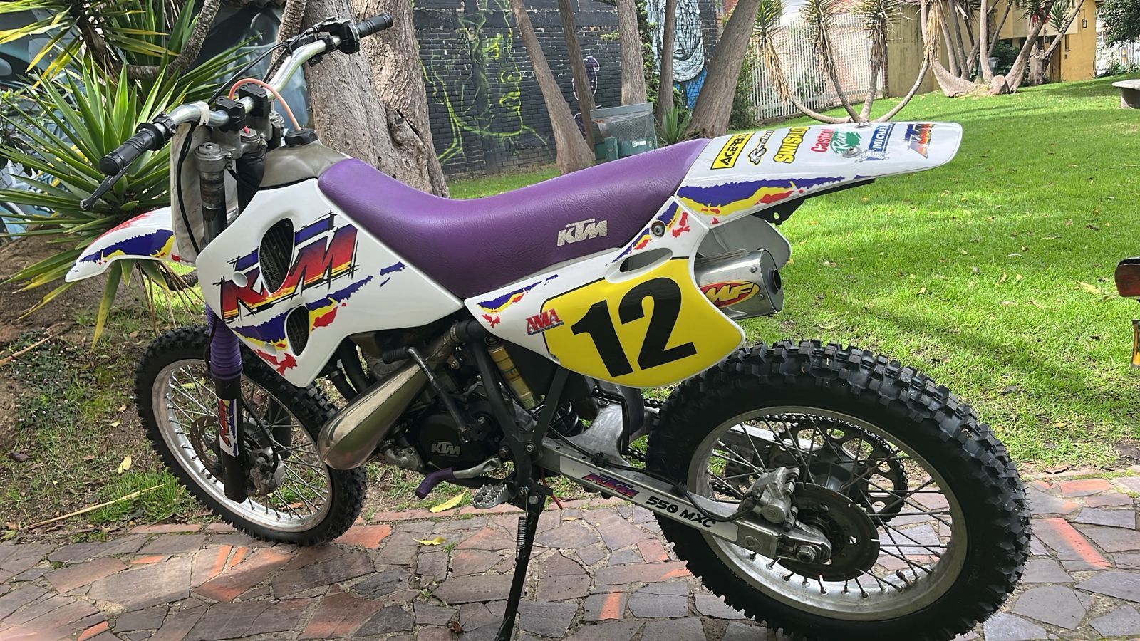 Moto todoterreno KTM morada y blanca con el número 12 estacionada sobre ladrillos al lado de césped verde.