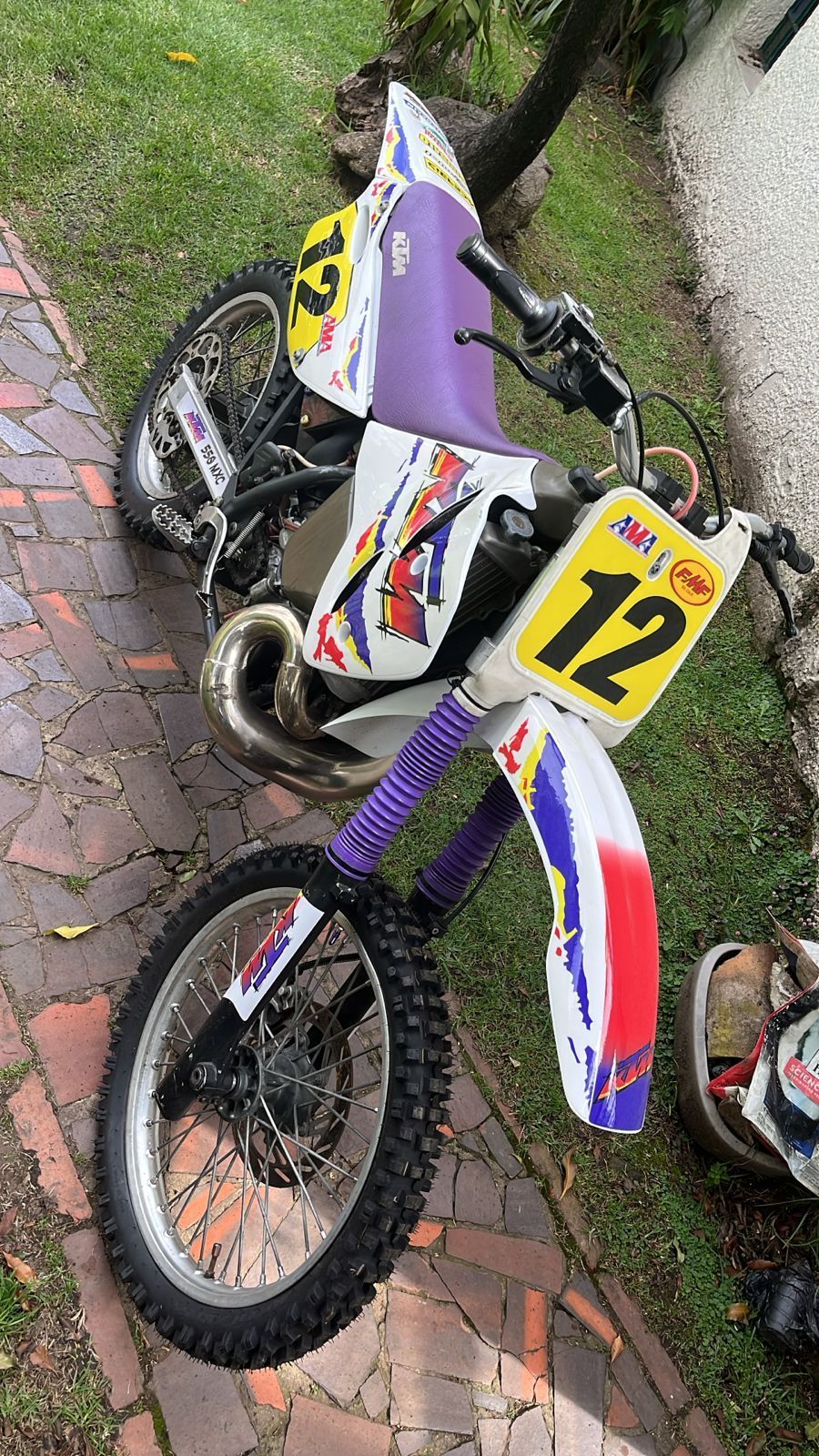 Moto todoterreno KTM de color blanco, morado y rojo, con el número 12, sobre una zona de césped.