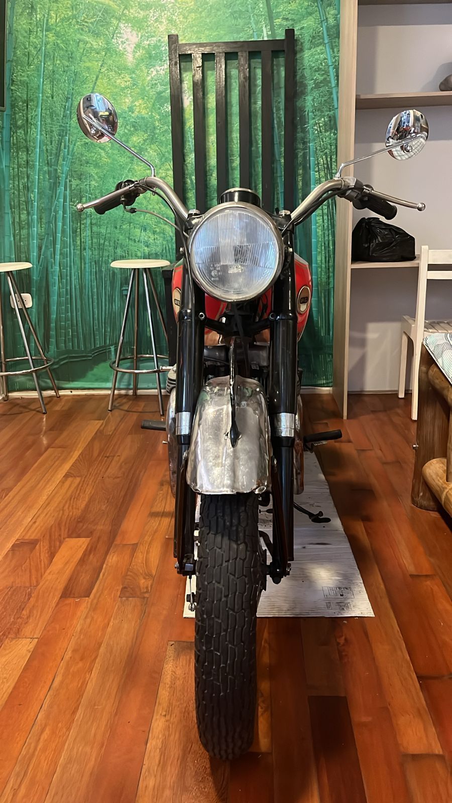 Motocicleta negra y roja, con faro redondo, estacionada sobre un piso de madera, frente a un mural forestal.