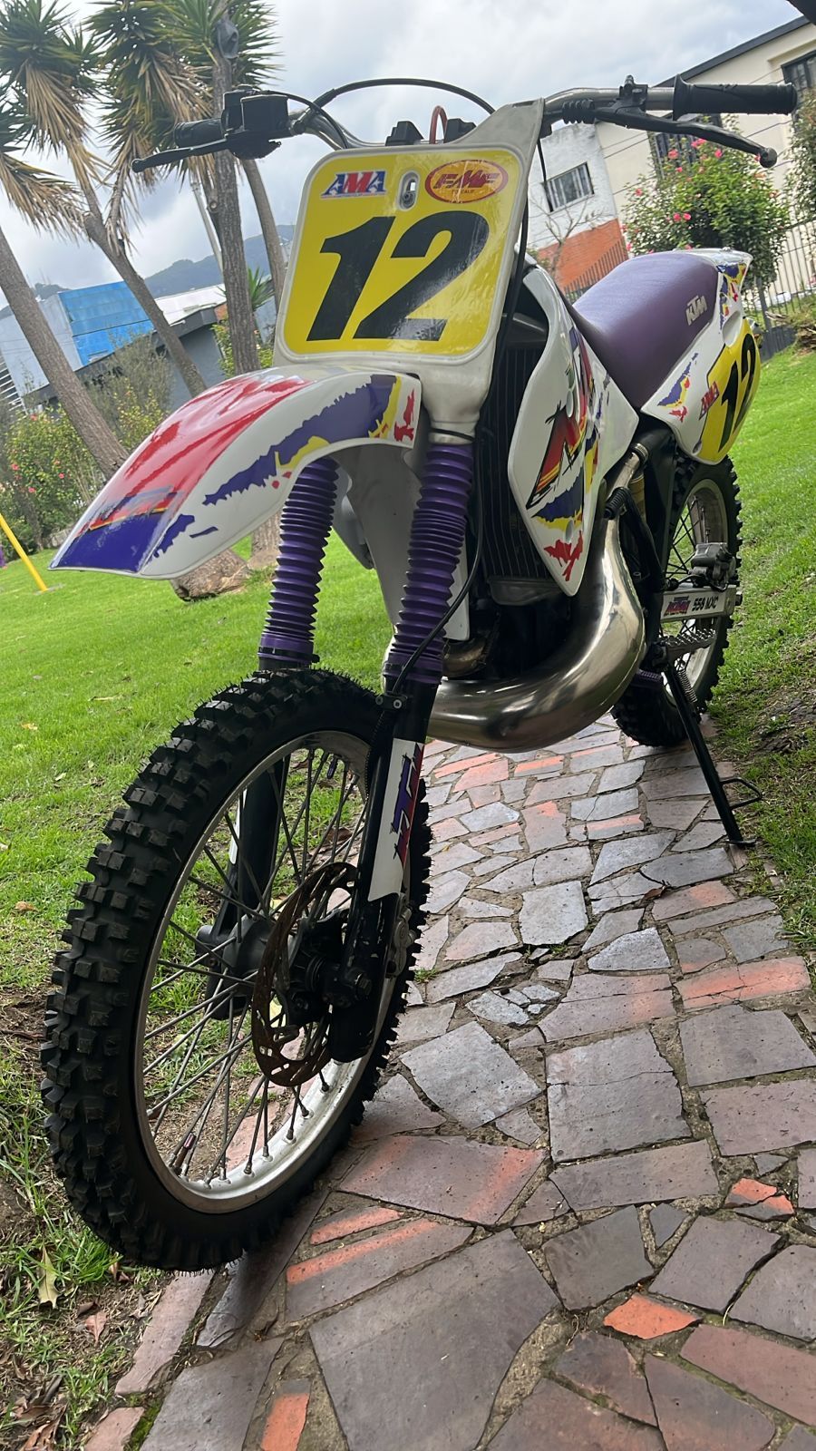 Una moto de motocross, número 12, estacionada en un camino de ladrillos, con suspensión violeta y carenado blanco y rojo.