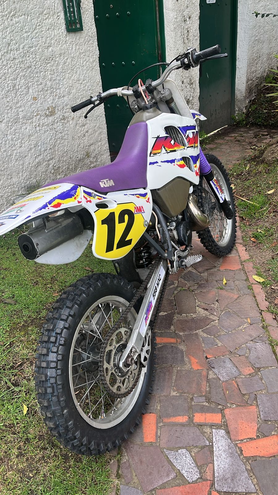 Moto de cross de color blanco, morado y amarillo estacionada en un camino de ladrillos con el número 12.