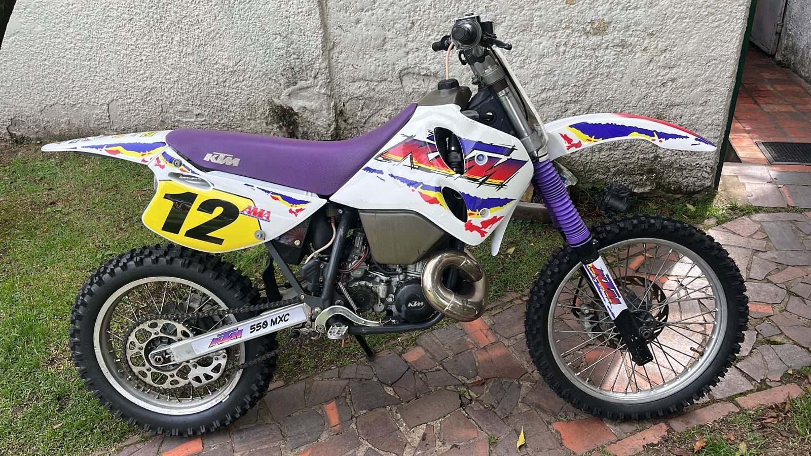 Moto todoterreno KTM con detalles en blanco, morado y amarillo. Número 12 en placa amarilla. Exterior.