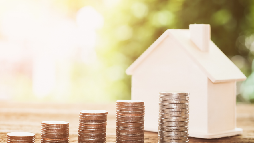 A model house is sitting next to stacks of coins on a wooden table.
