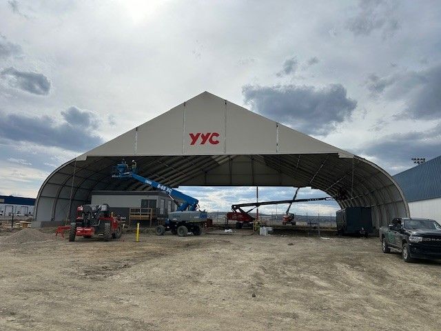 Large white YVC tent structure at a dirt airfield with vehicles and aircraft underneath, under a cloudy sky