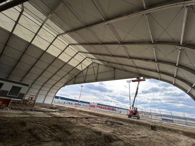 Large arched hangar interior with dirt floor and a small crane near the open side under a cloudy sky