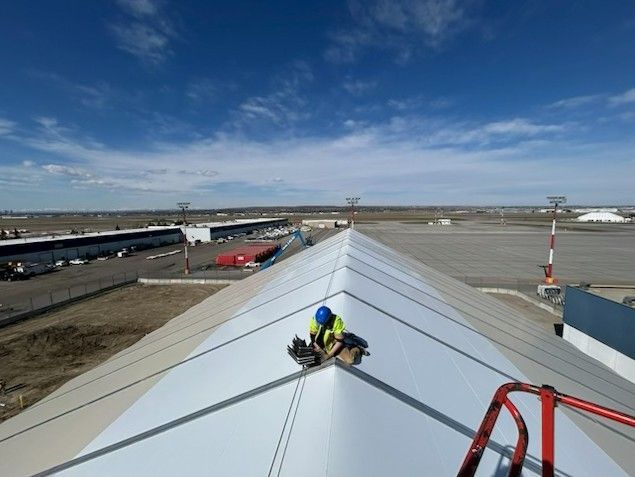 Worker kneeling on a white rooftop beside a red railing under a clear blue sky