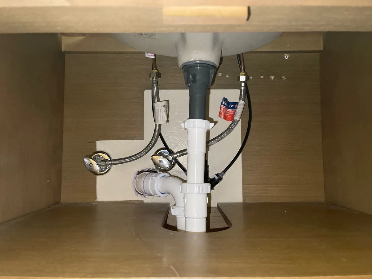 Under-sink plumbing with pipes, shutoff valves, and a garbage disposal inside a wooden cabinet