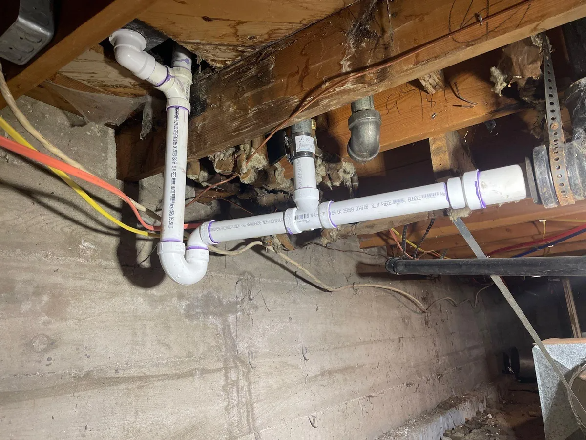 Exposed white PVC pipes and fittings under wooden floor joists in a basement crawl space.
