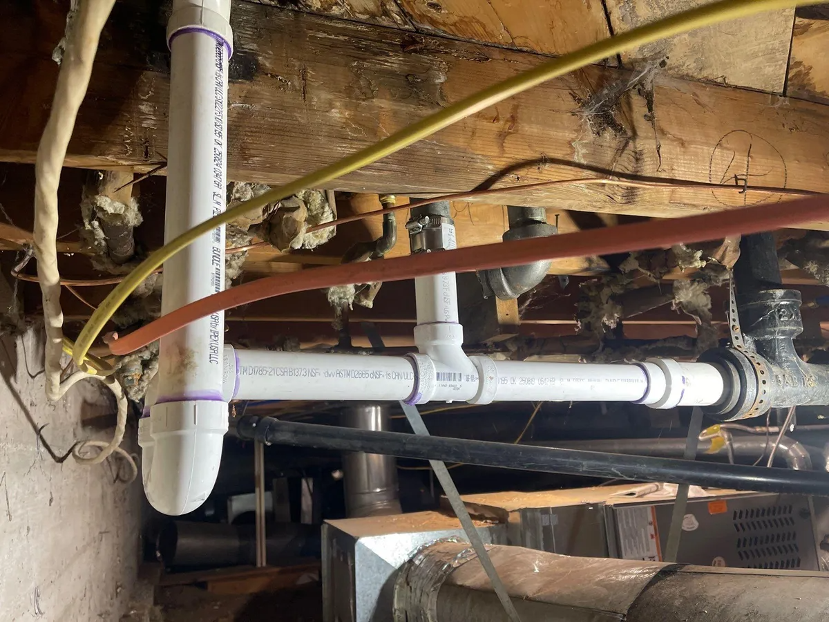 Exposed PVC pipes and wiring under a basement floor, with a wall opening and wooden beams.