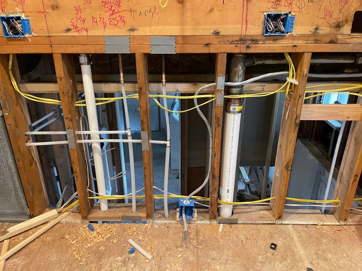 Exposed wall studs with plumbing pipes and electrical wiring in an unfinished room, with a subfloor opening.