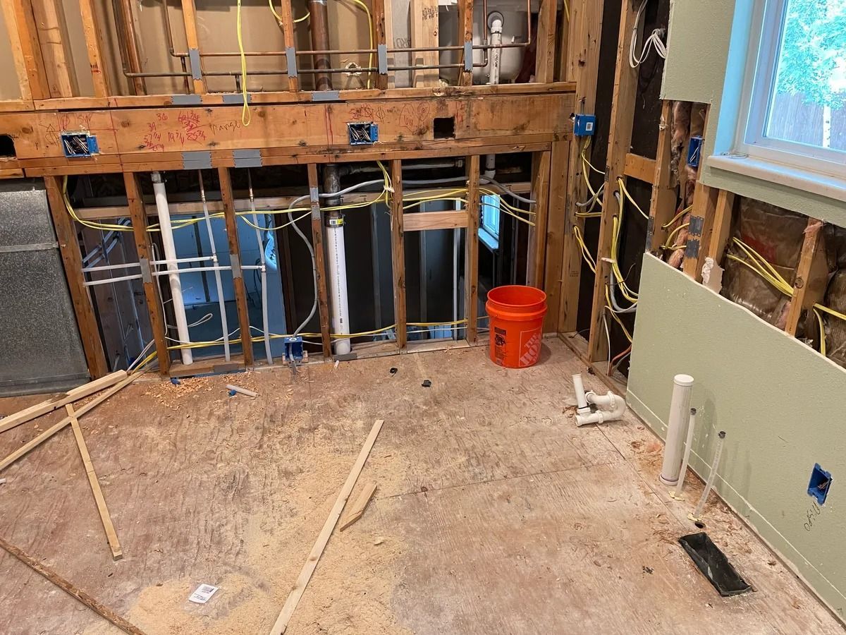 Renovated room with exposed studs, unfinished flooring, pipes, and an orange bucket near a window