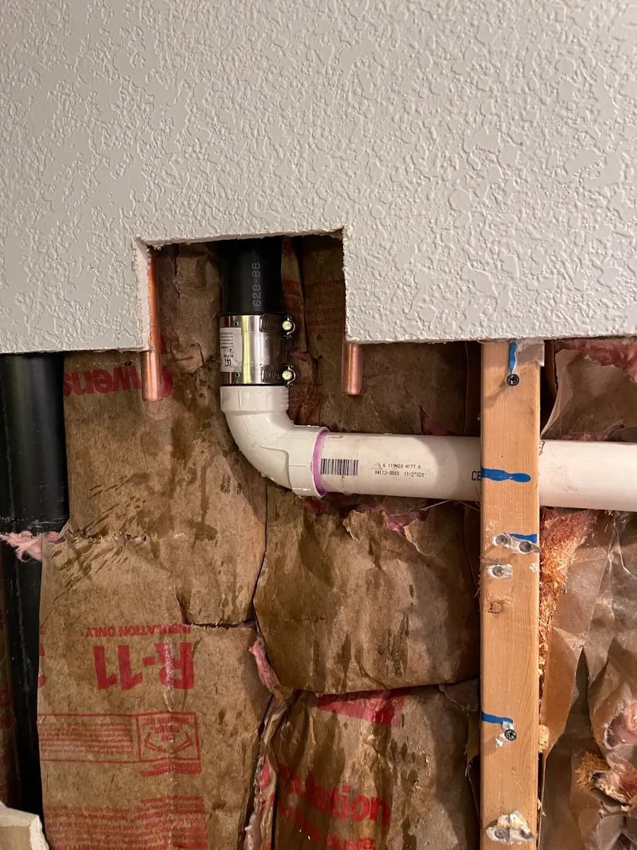 Open wall exposing white PVC plumbing elbow and pipe amid pink insulation and exposed framing.