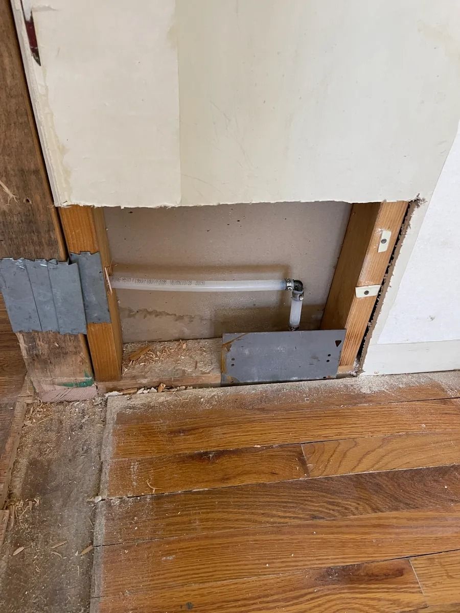 Damaged wall panel removed, exposing pipes and a floor opening above hardwood flooring.