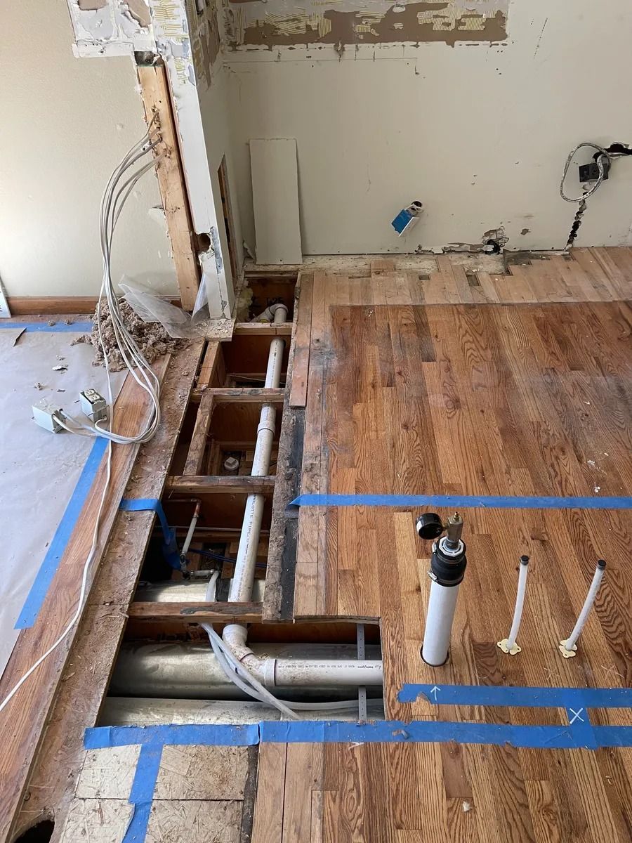 Partial flooring removed in a room, exposing joists, wiring, and a white pipe during renovation