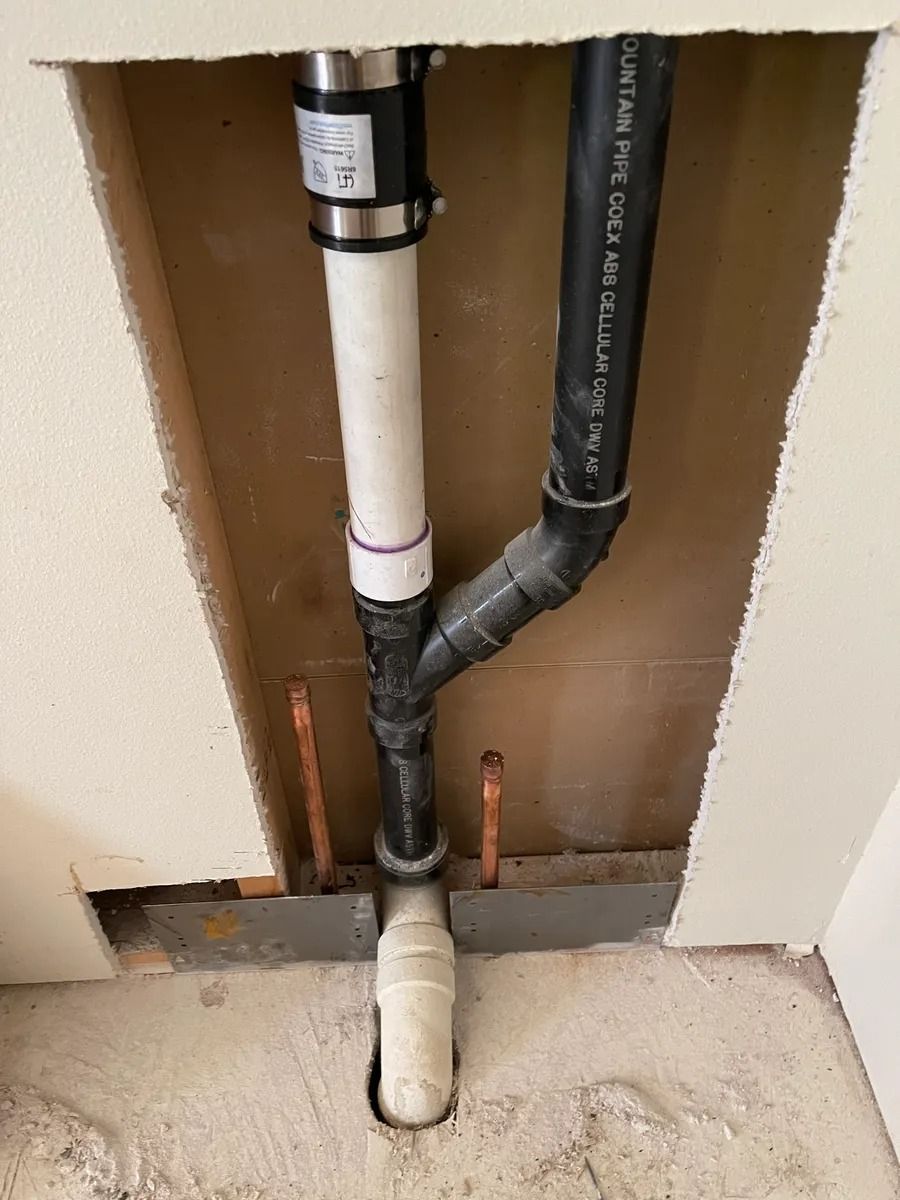 Exposed plumbing pipes inside a wall opening, with white and black drain lines and a floor vent stub.