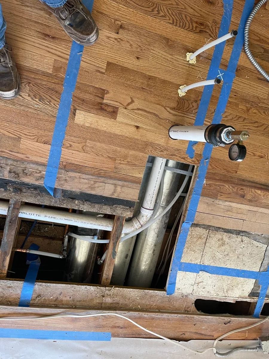Exposed plumbing and ductwork through a cutout in subflooring, with blue tape marking the area.