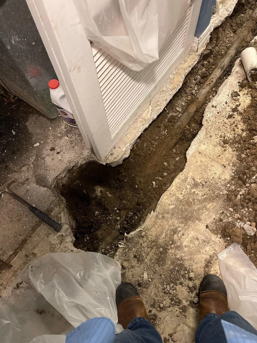Clogged drainage trench beside a building doorway, with muddy debris and boots visible at the bottom.