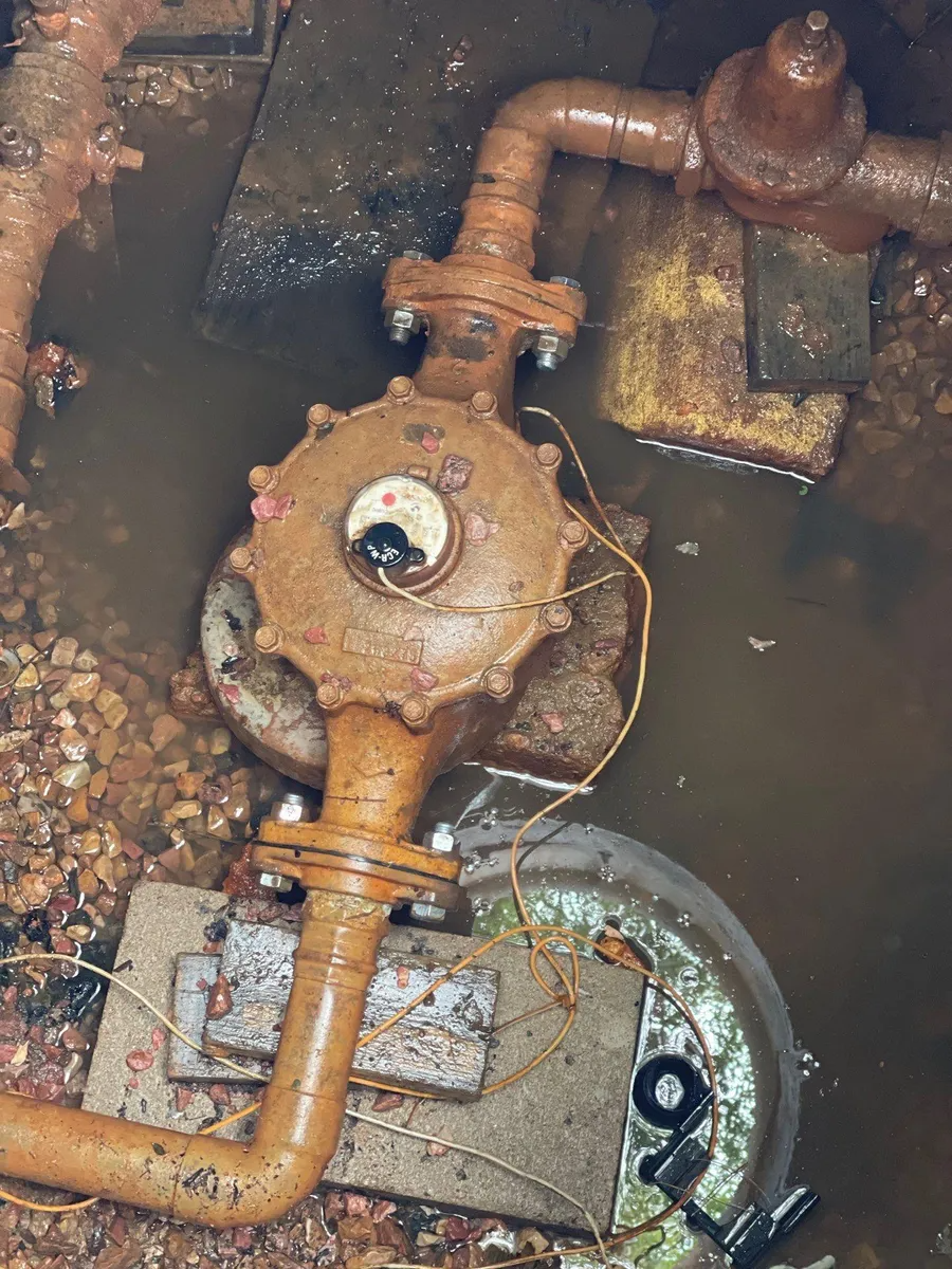 Rusty water valve and pipes in a muddy pit, with standing water and gravel around them