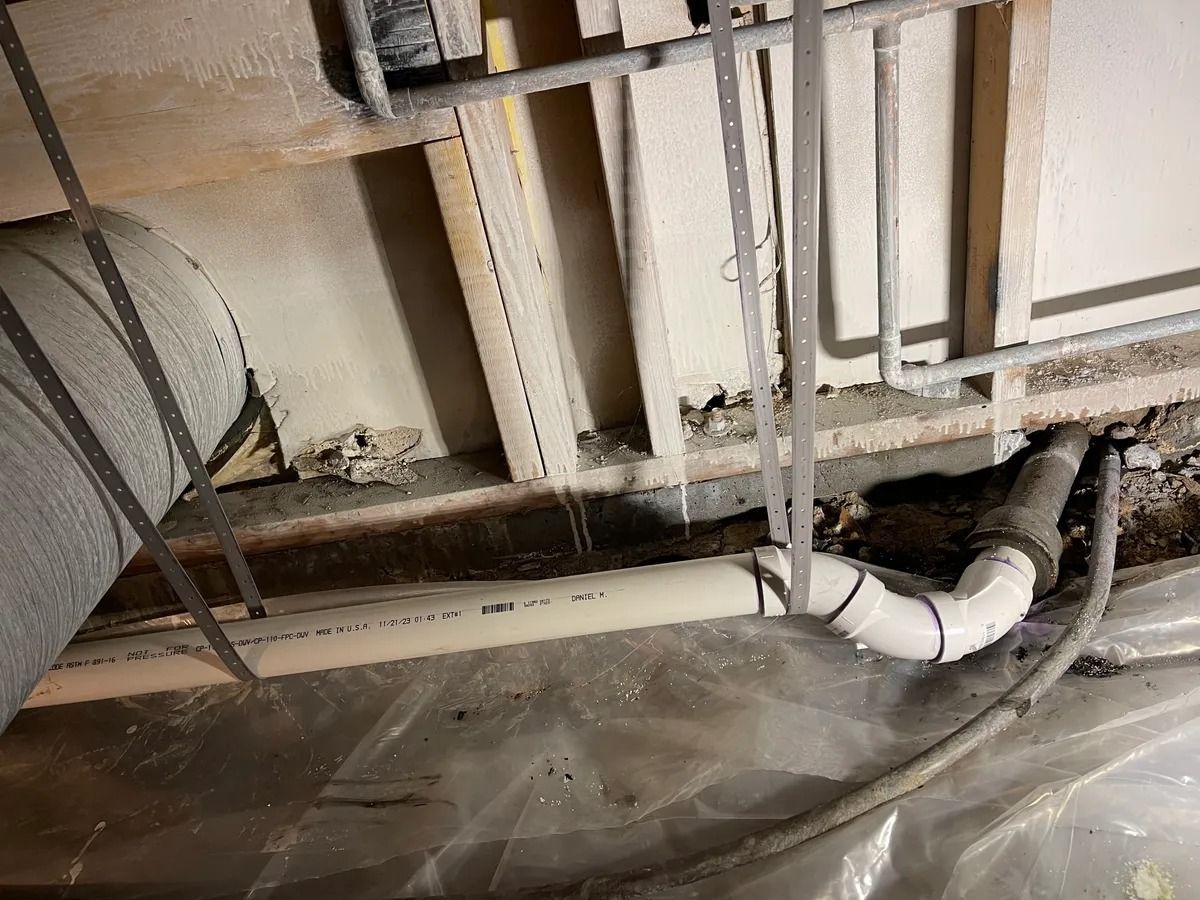 Plumbing under a damaged floor with exposed pipes, insulation, and plastic sheeting on the ground