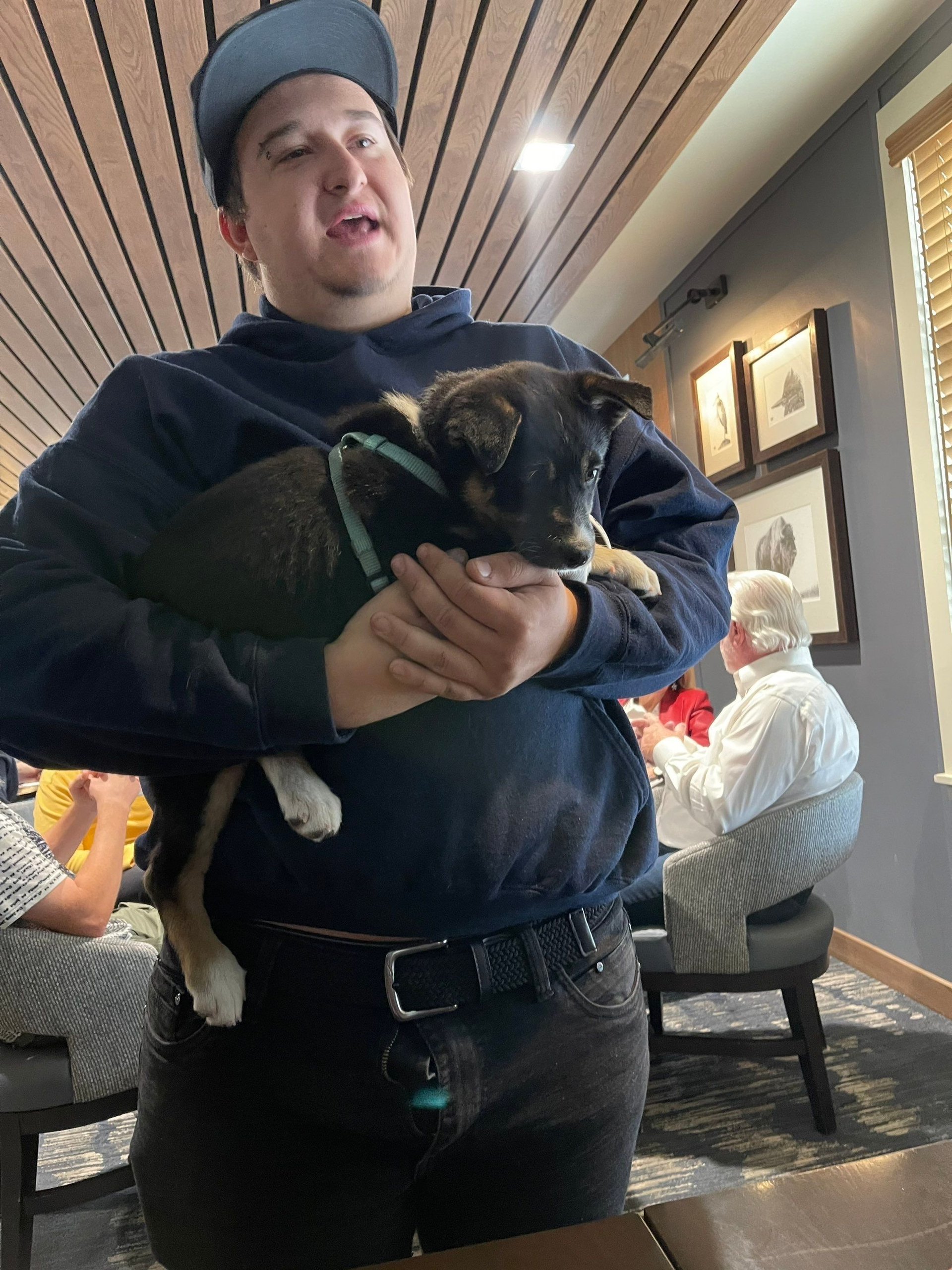 Person holding a small black-and-tan dog indoors, with people seated in the background.