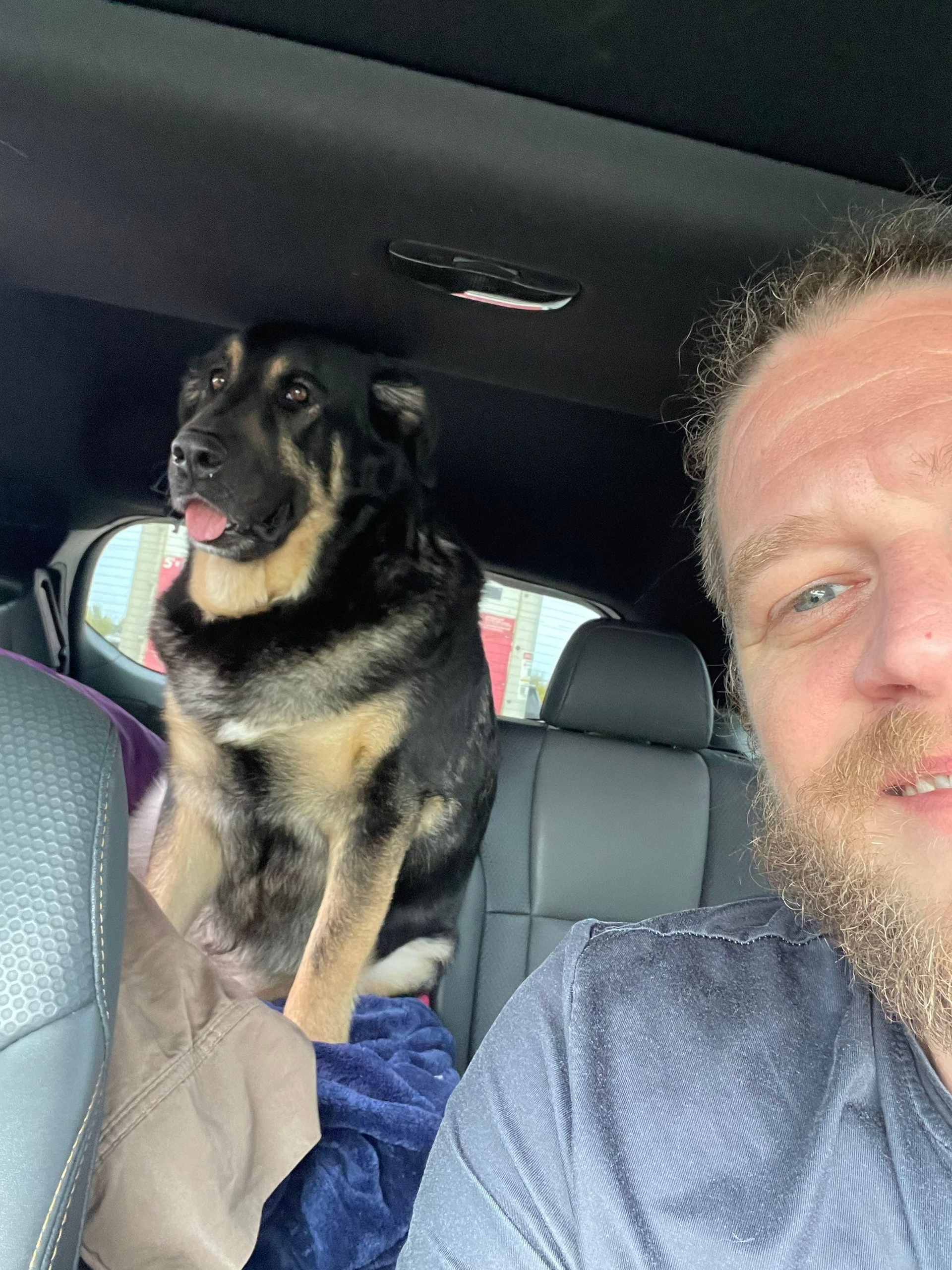 Dog sitting on a car seat beside a man taking a selfie.