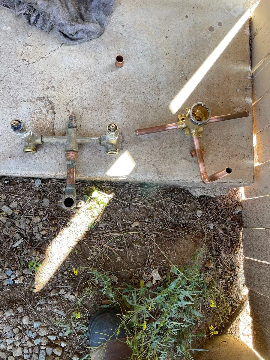 Rusty metal valve assembly on a concrete slab, with sunlight and weeds below.