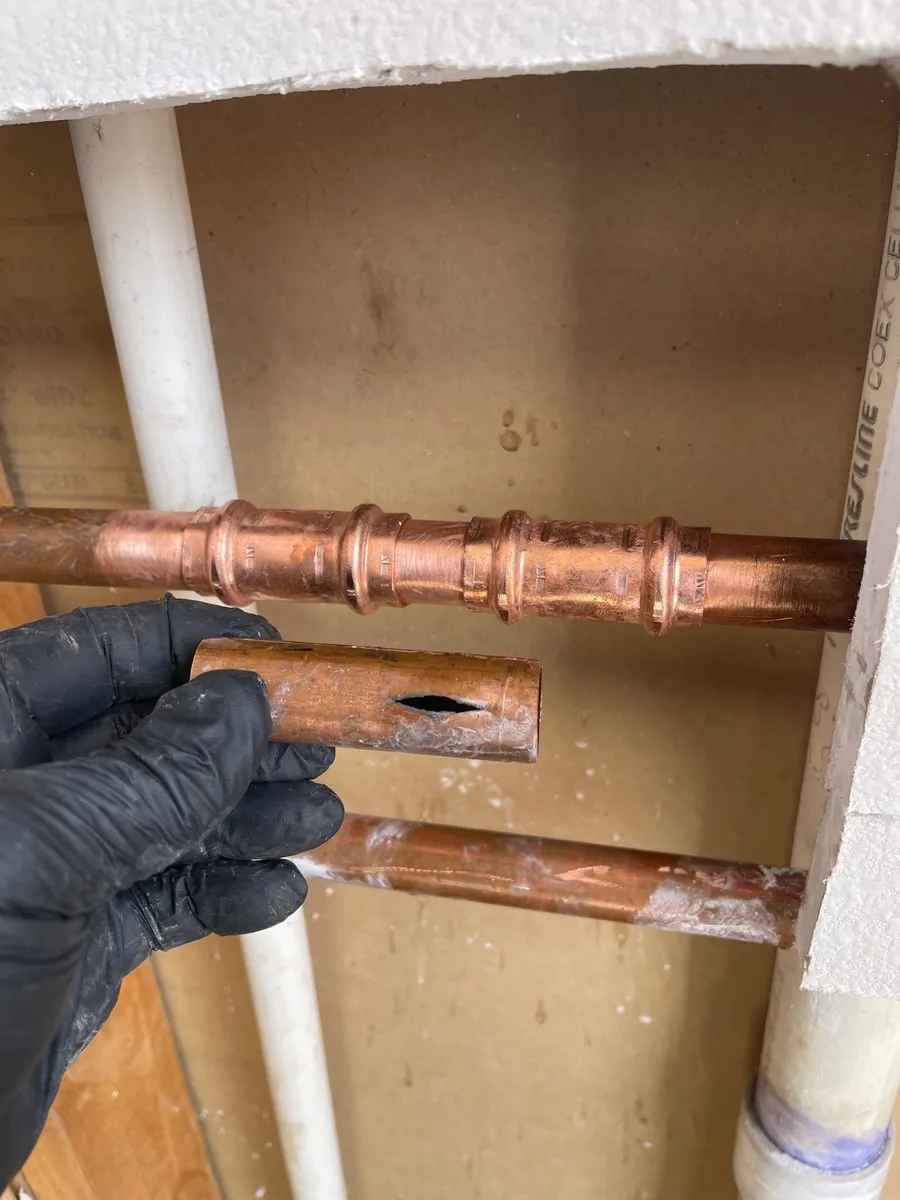 Gloved hand holding a copper pipe fitting inside an open wall cavity with exposed plumbing.