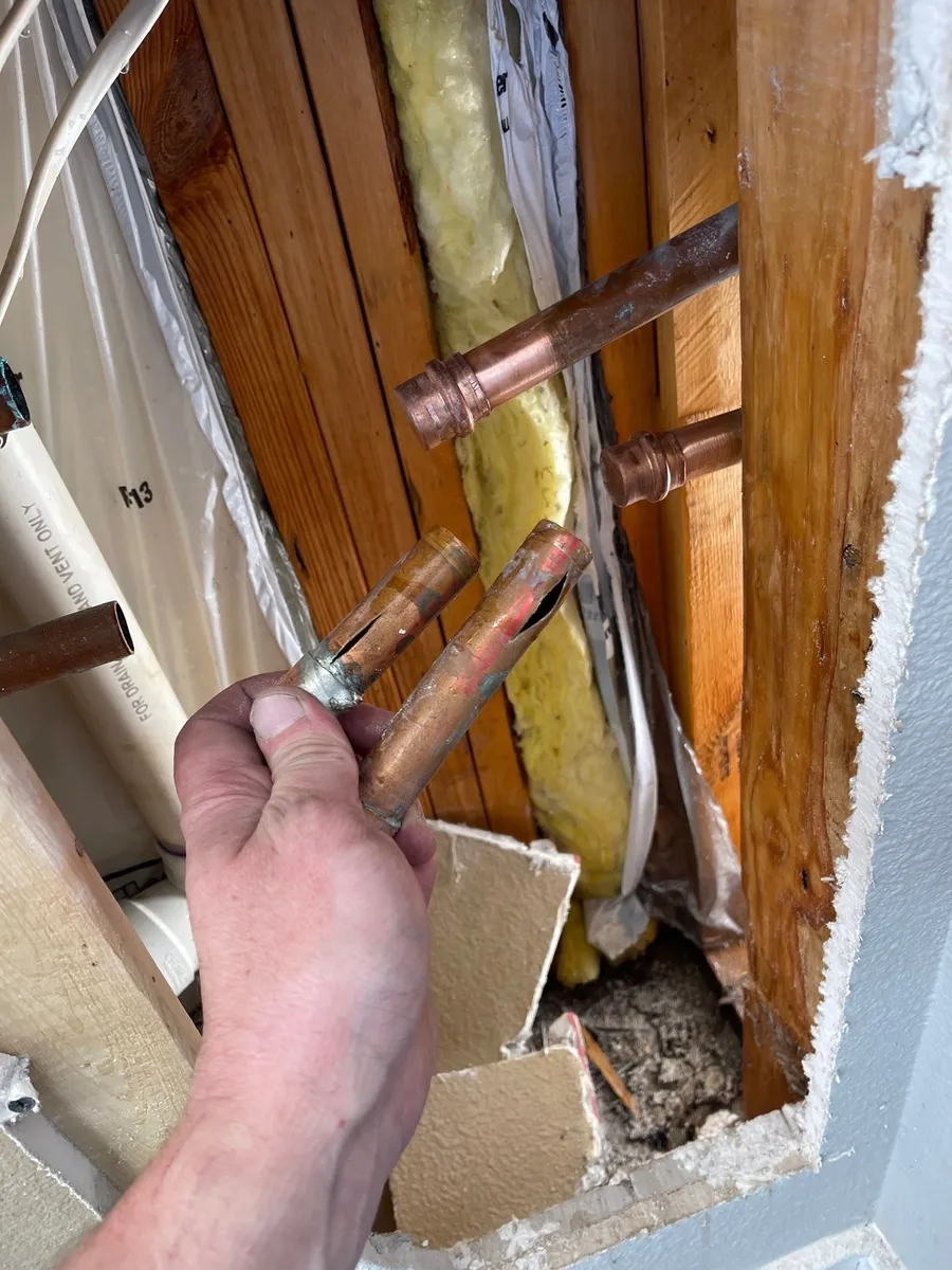 Hand holding copper pipes inside a wall cavity with insulation and drywall debris