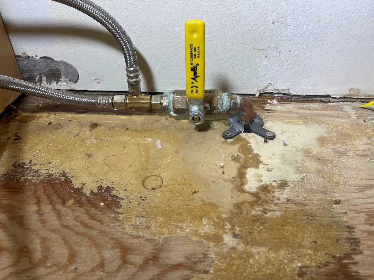 Leaking pipe with yellow shutoff valve and water damage on a floor against a wall
