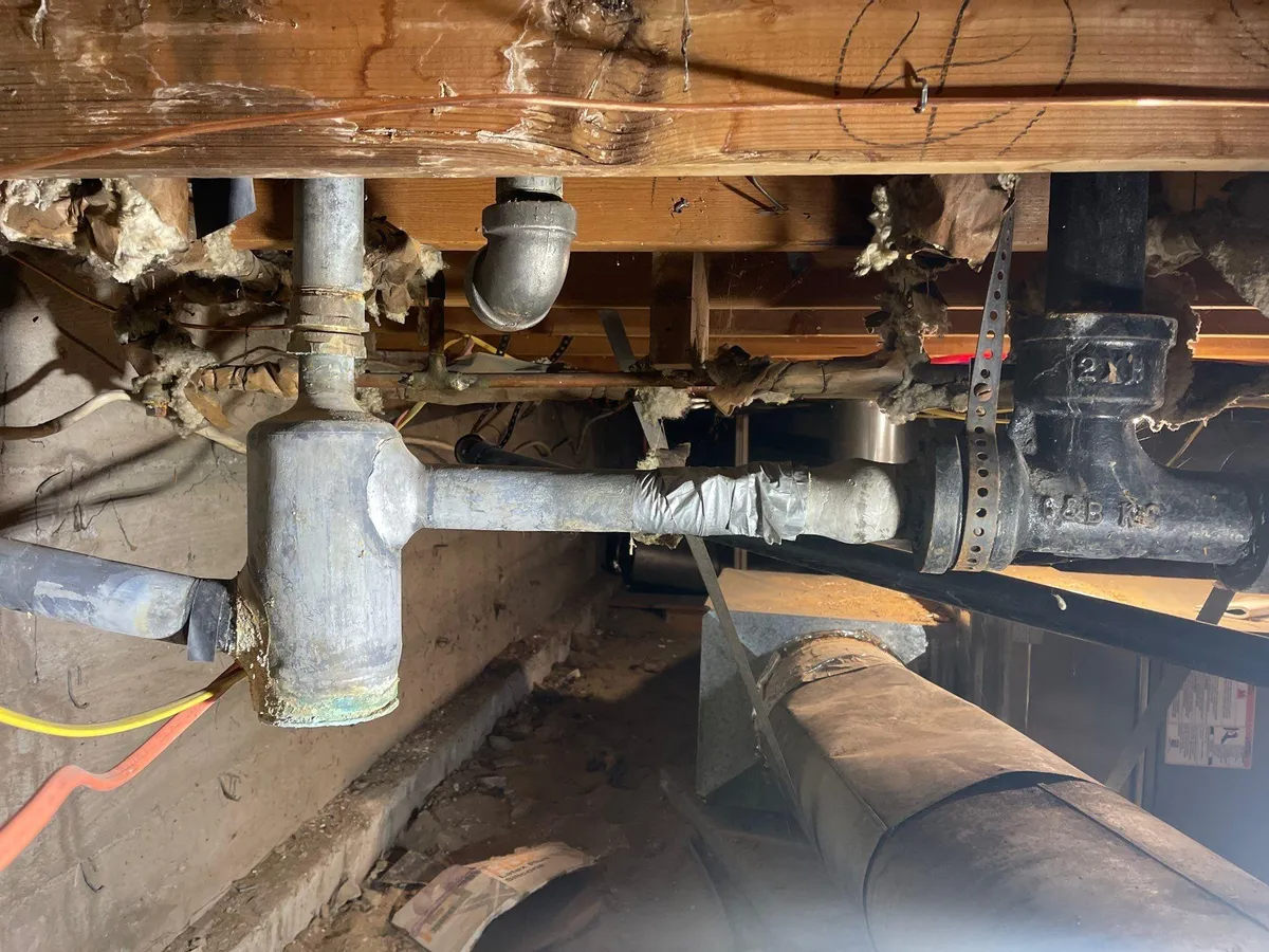 Close-up of exposed pipes and fittings under a wooden floor, showing a basement plumbing repair area.