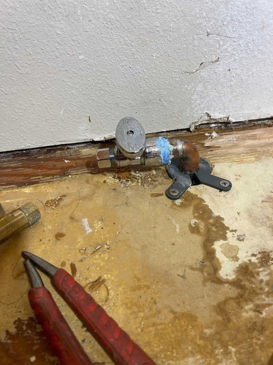 Blue faucet leaking under a wall, with wet floor and red-handled pliers nearby