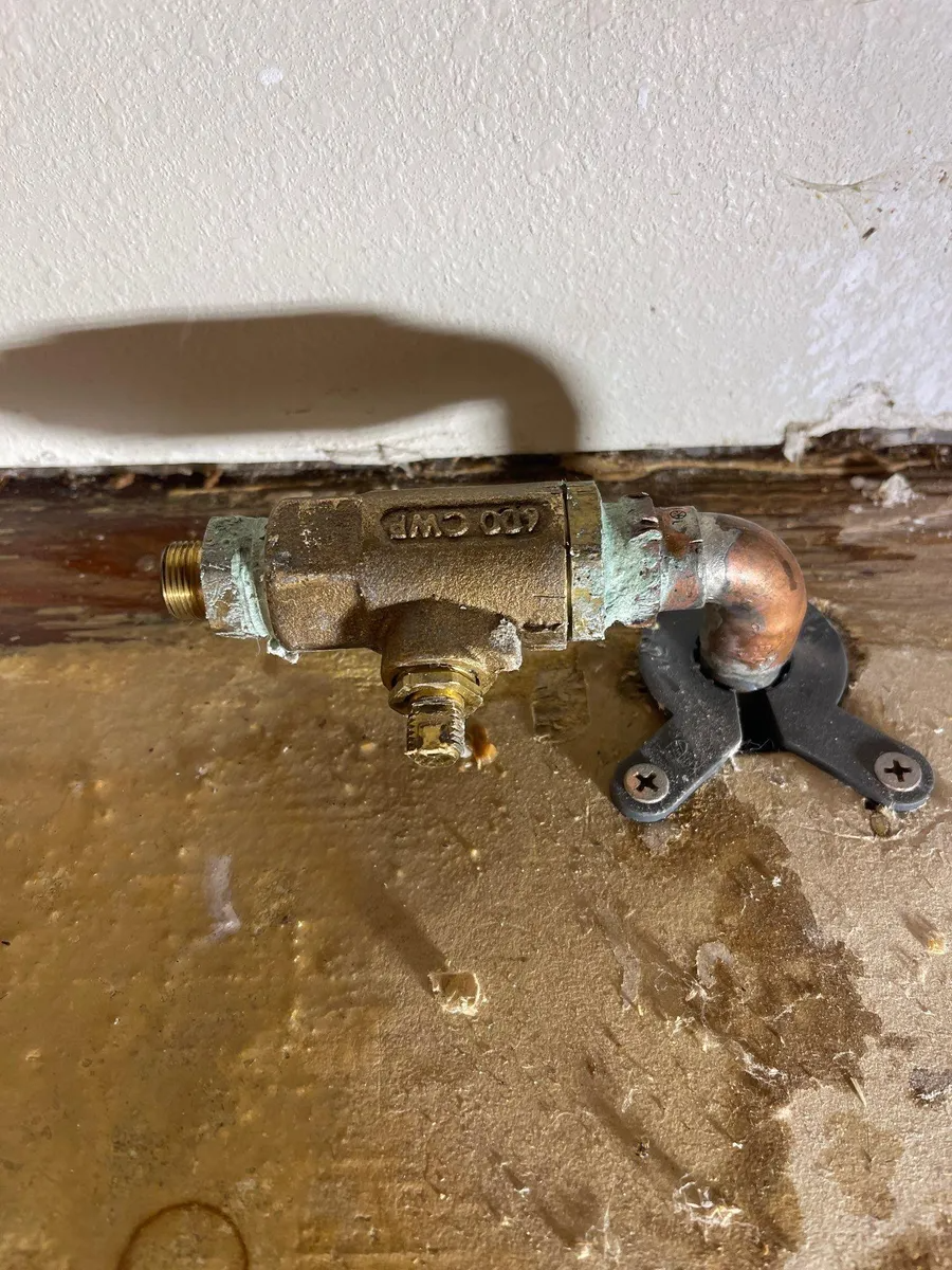 Corroded brass valve and pipe fittings mounted near a wall on a dirty floor.