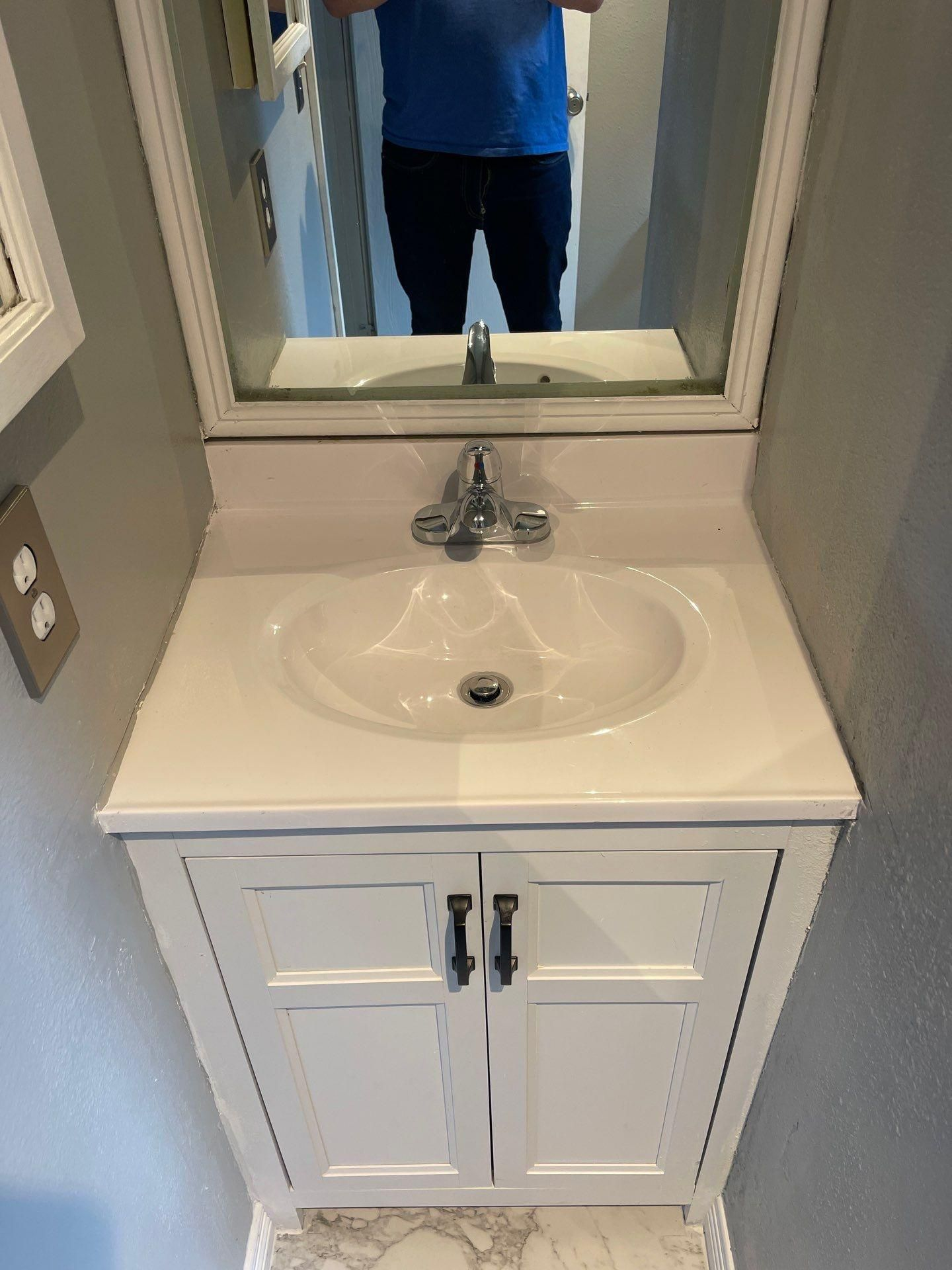 White bathroom vanity with sink and mirror; person in blue shirt reflected in mirror.