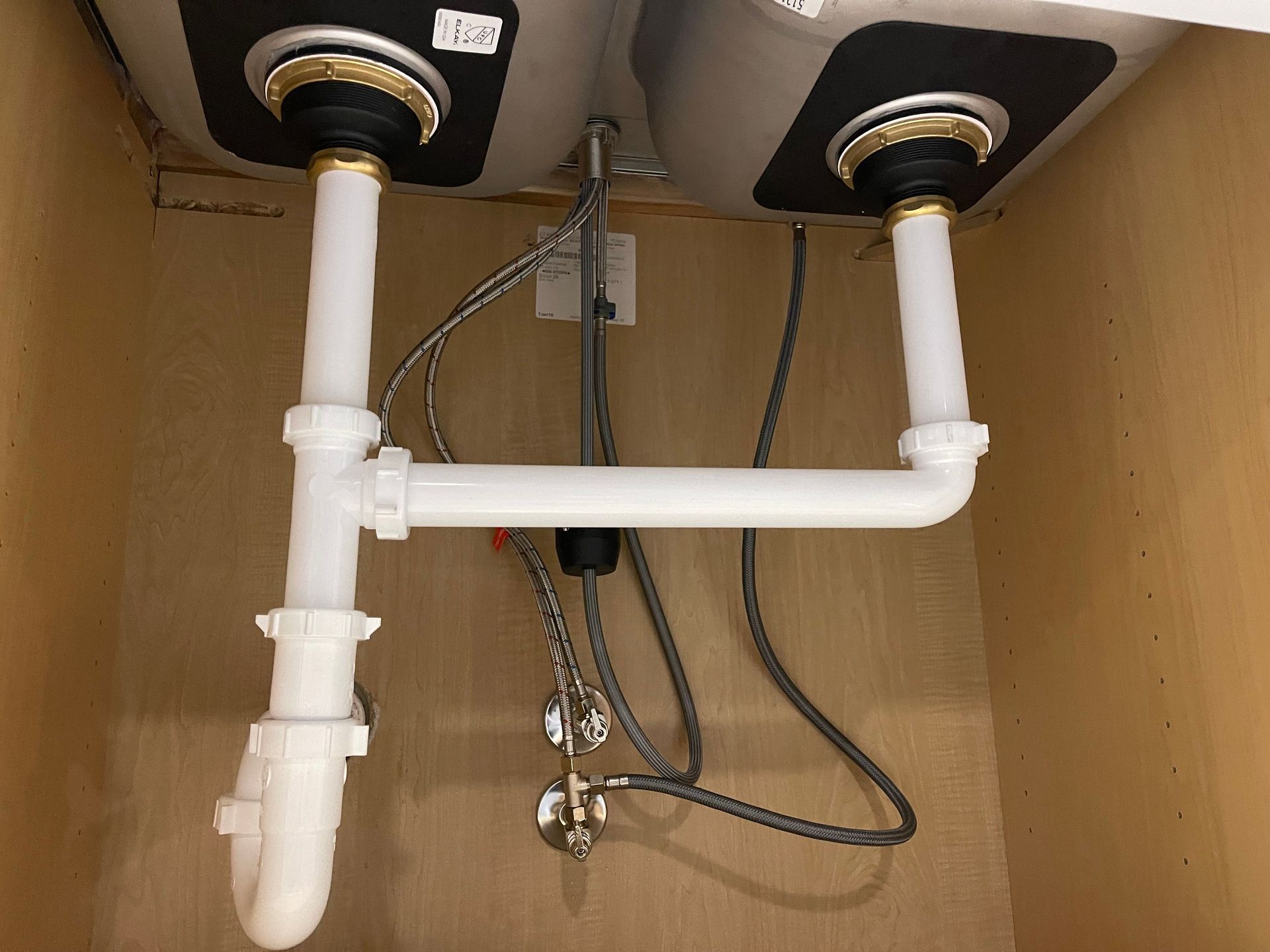 Under-sink plumbing with two drain pipes, white PVC trap, and visible flexible hoses in a cabinet