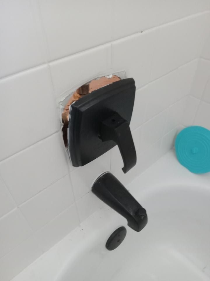 Black wall-mounted bathtub faucet with detached handle, exposing copper piping against white tile.
