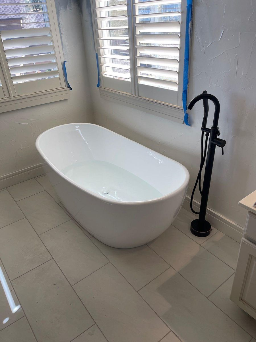 Modern freestanding white bathtub beside a black floor-mounted faucet in a bright tiled bathroom