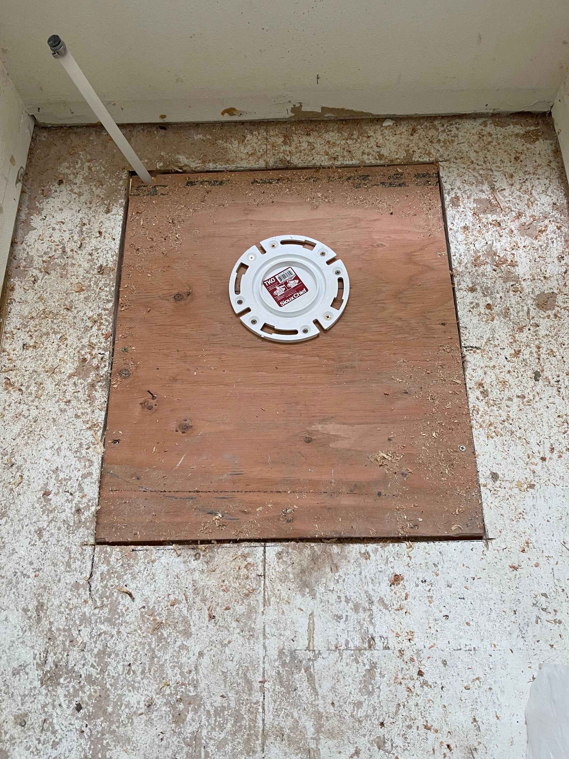 Wooden board with a circular drain cover in a concrete corner, with a pipe at the left.