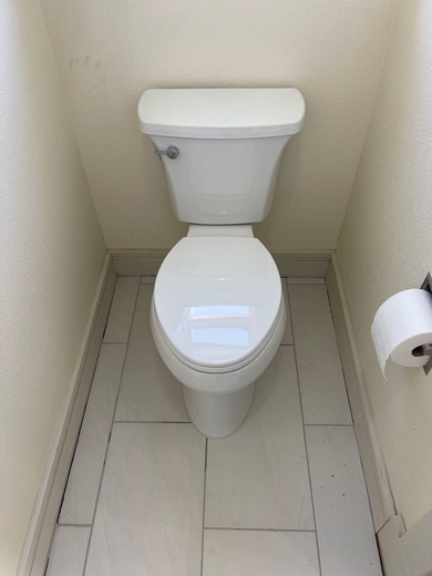 Small white toilet in a narrow beige bathroom stall, with toilet paper on the right wall.